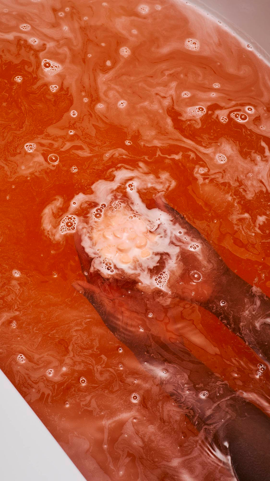 A vibrant coral-coloured "Comforter Rose" bath bomb held by two hands just beneath the surface of orange, foamy bathwater. The bath bomb is partially covered in thick white bubbles, which swirl and spread across the warm-toned water.