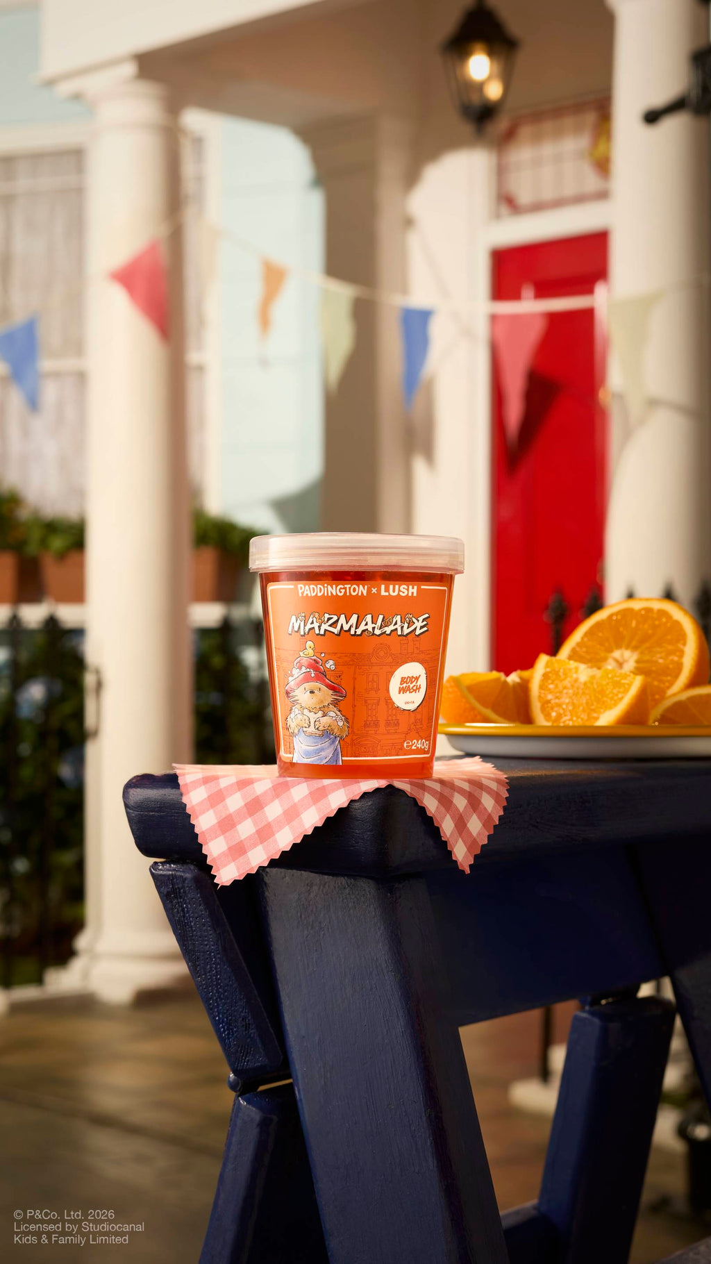 A clear pot of 'Marmalade' body wash from the Paddington x Lush range sits on a blue wooden bench outdoors. The scene includes bright sunshine, a red front door, bunting, and fresh orange slices on a gingham cloth.